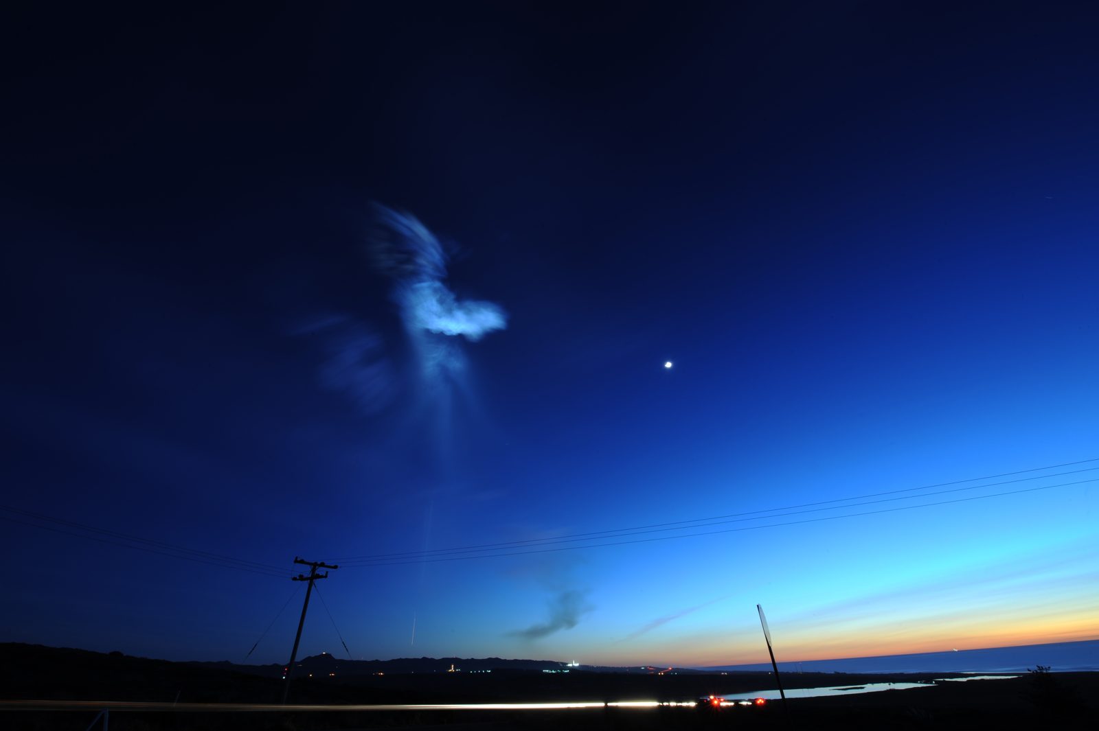A pale blue, feather-shaped rocket exhaust cloud glowing high in a deep-blue twilight sky above a dark coastal horizon, with a bright planet to its right.
