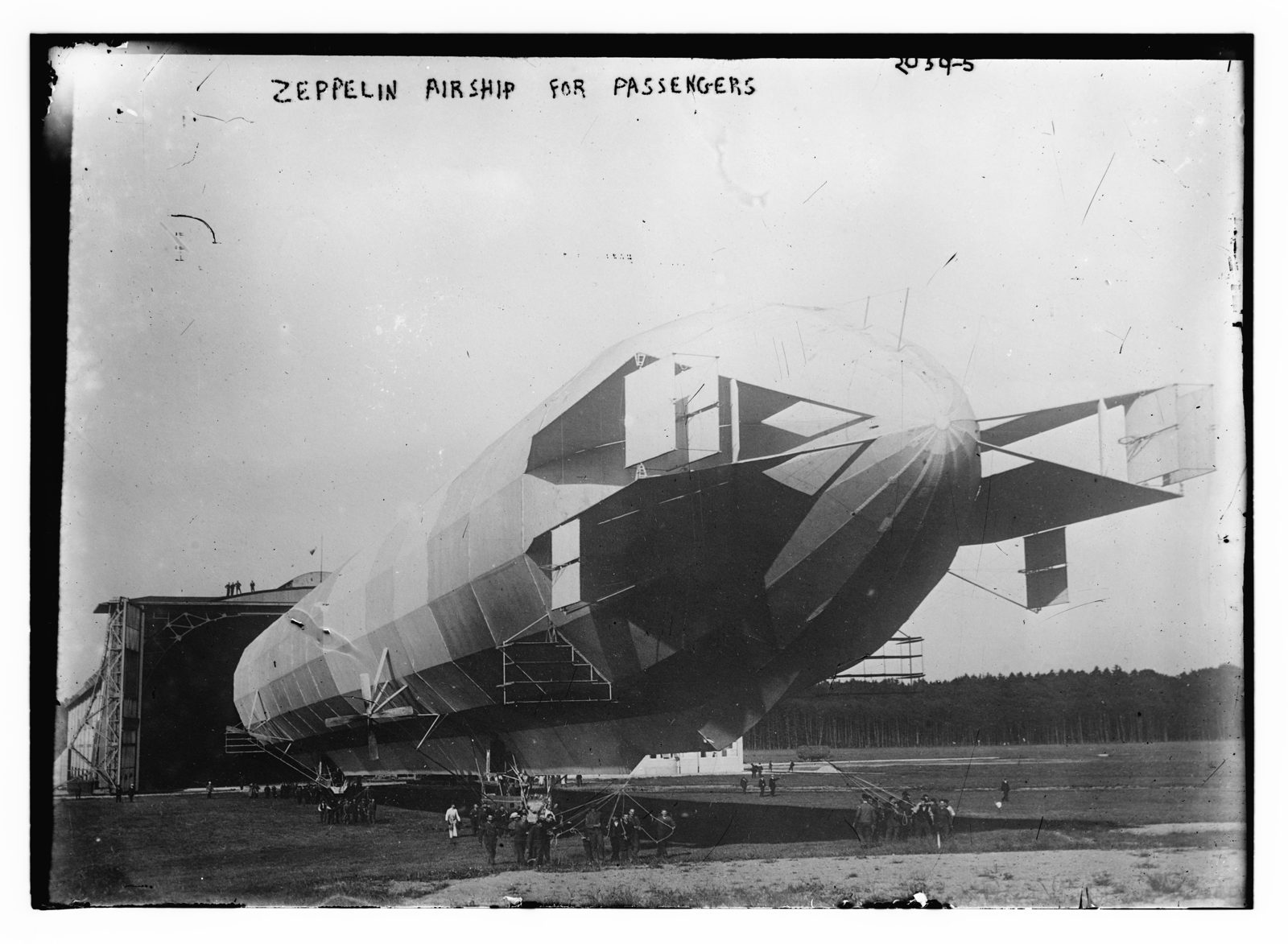Black-and-white photo of a large rigid Zeppelin airship on the ground beside its hangar, with small figures of people standing beneath it; handwritten caption 'Zeppelin airship for passengers.'
