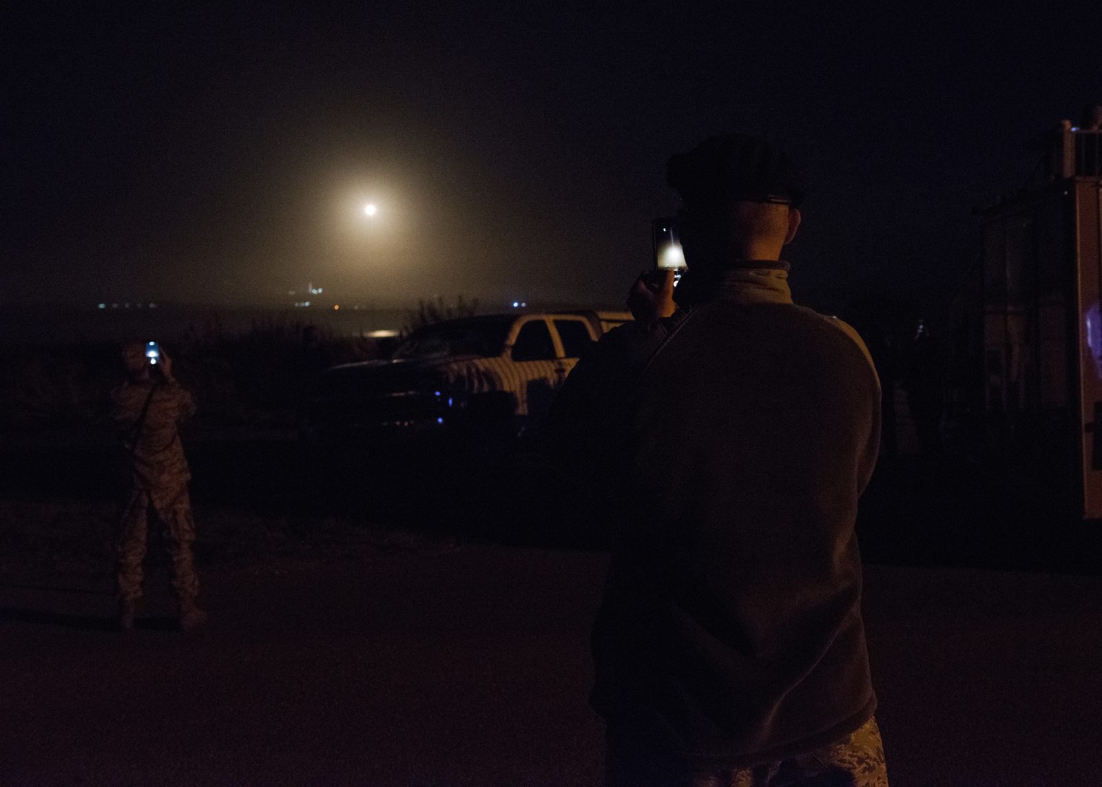 At night, two people, one in camouflage uniform, hold up phones to film a bright glowing point of light rising in a hazy sky over the coast, with a pickup truck in the dark foreground.
